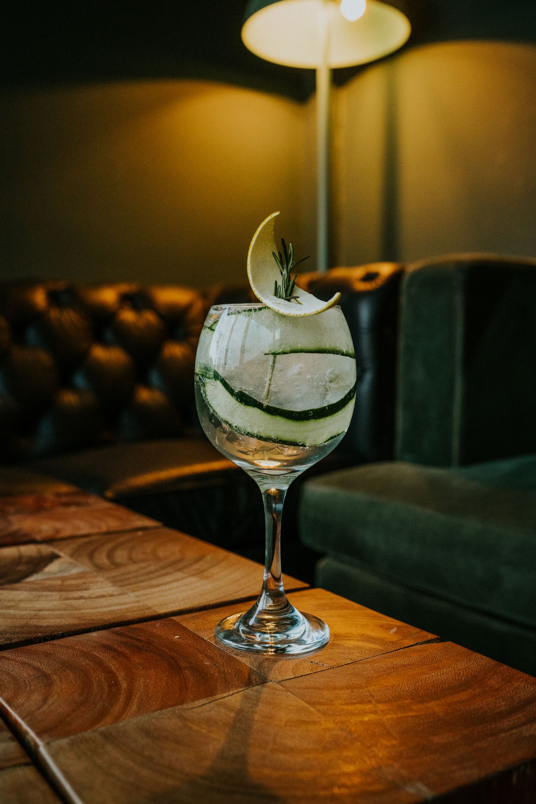 Aesthetic shot of a gin and tonic cocktail with cucumber and lemon garnish in a dimly lit cozy bar.