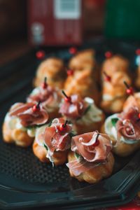 Many tasty profiteroles decorated with fresh prosciutto and skewers served on black tray on terrace with building against blurred background