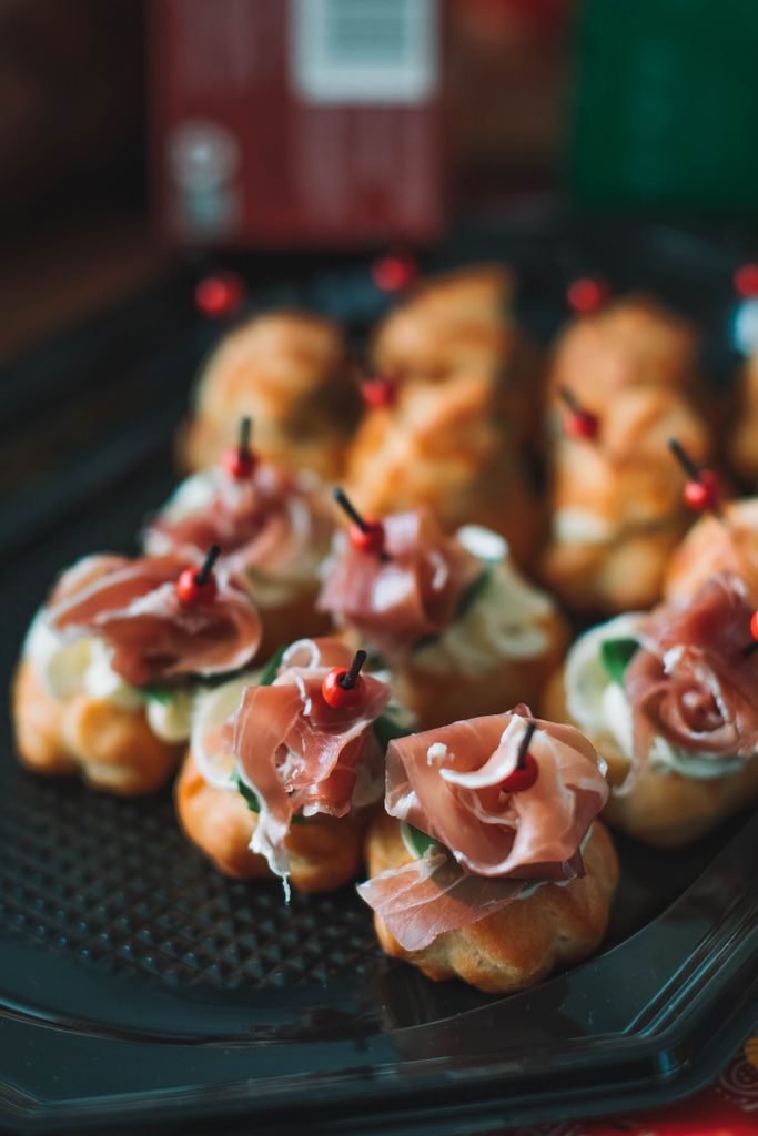 Many tasty profiteroles decorated with fresh prosciutto and skewers served on black tray on terrace with building against blurred background
