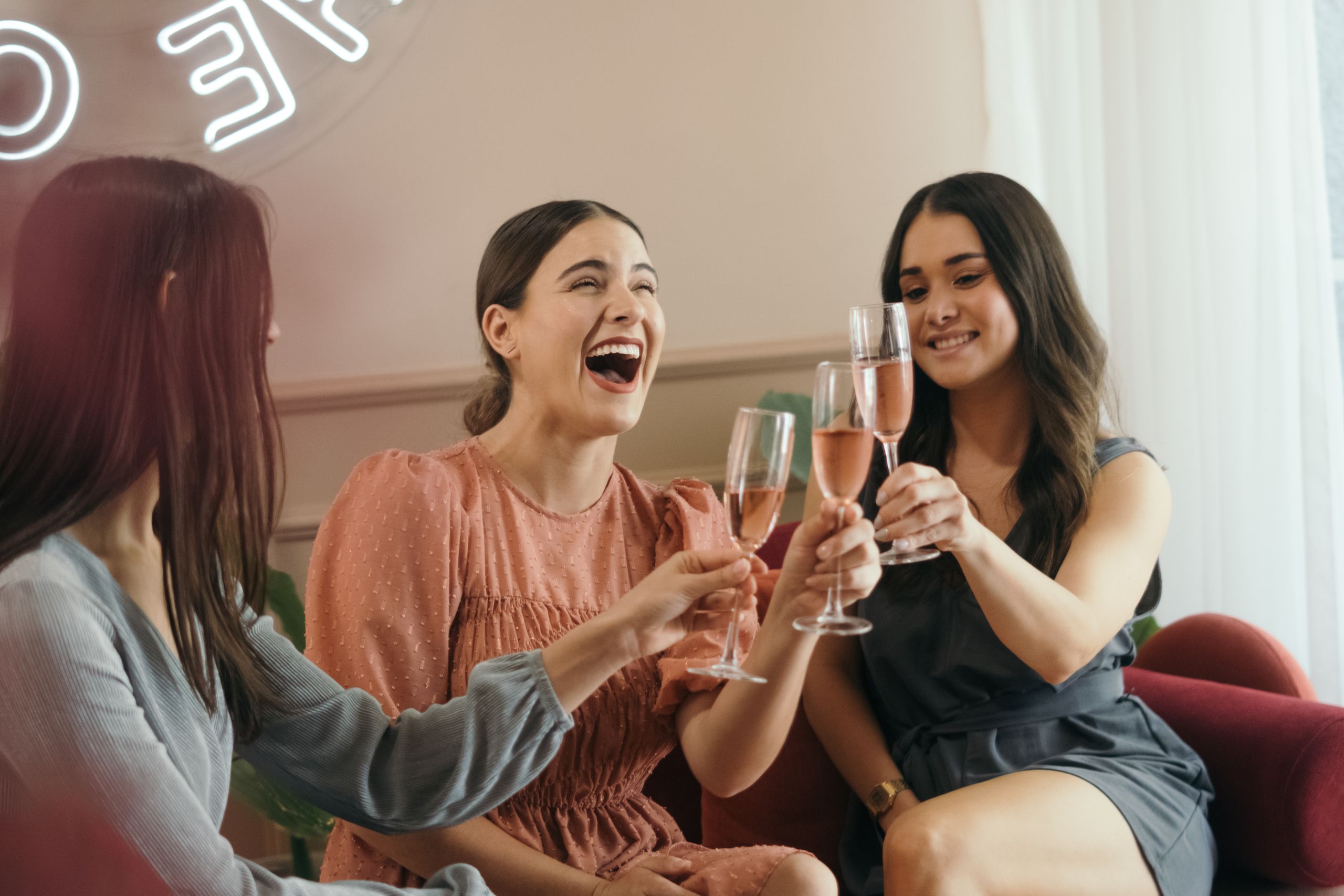 Three women laughing and clinking champagne glasses in a joyful indoor celebration.